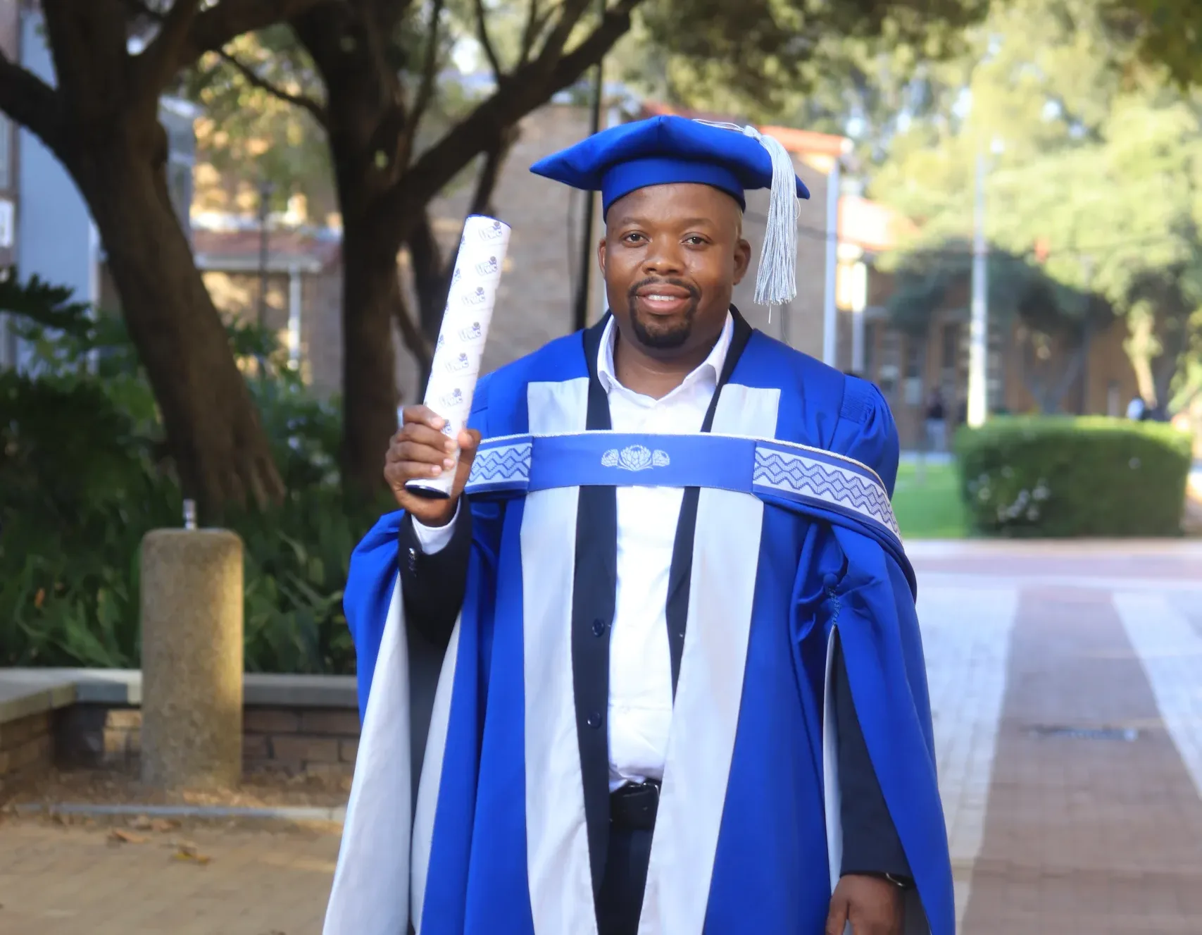 DR Jacob Thamara – Vaal University of Technology Graduate in a royal blue cap and gown holding a diploma on a campus walkway.