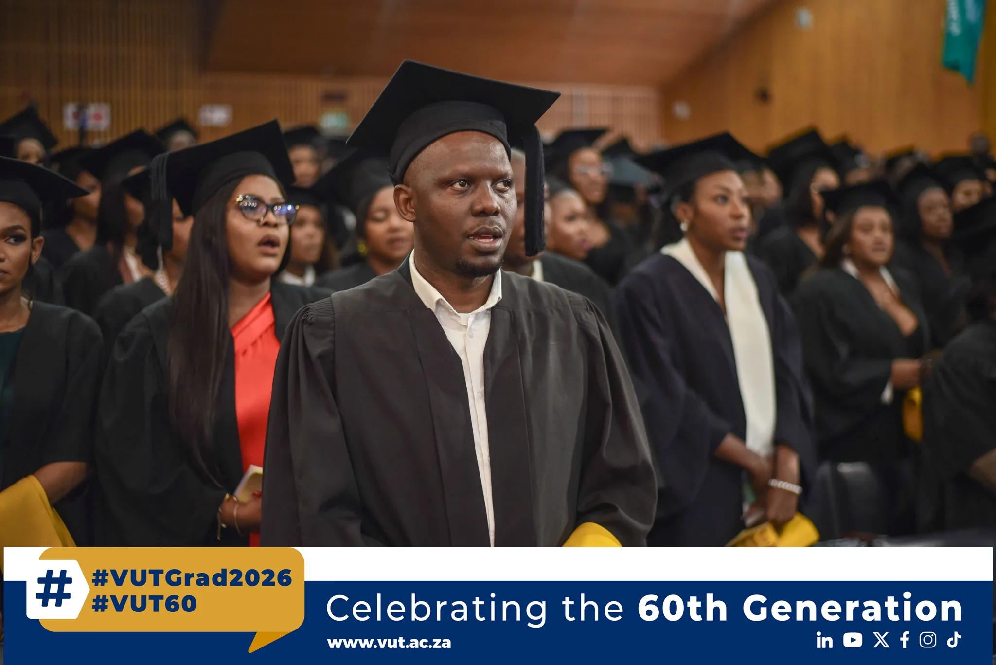 Graduate in cap and gown stands among fellow graduates during a ceremony, with a banner reading #VUTGrad2026 at the bottom.