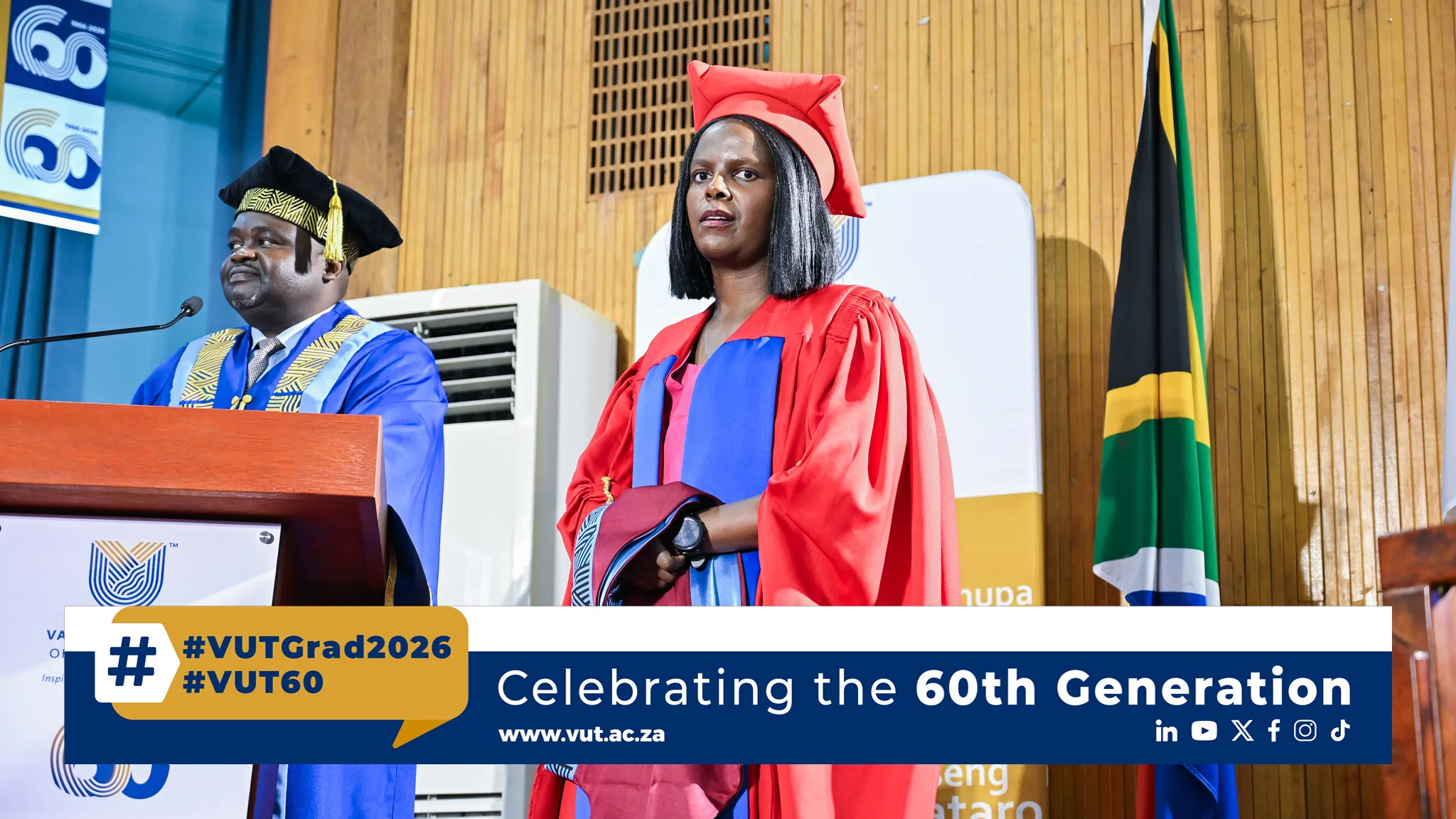 Graduate in red gown and cap stands beside man in blue gown at a podium during a ceremony; SA flag in background.