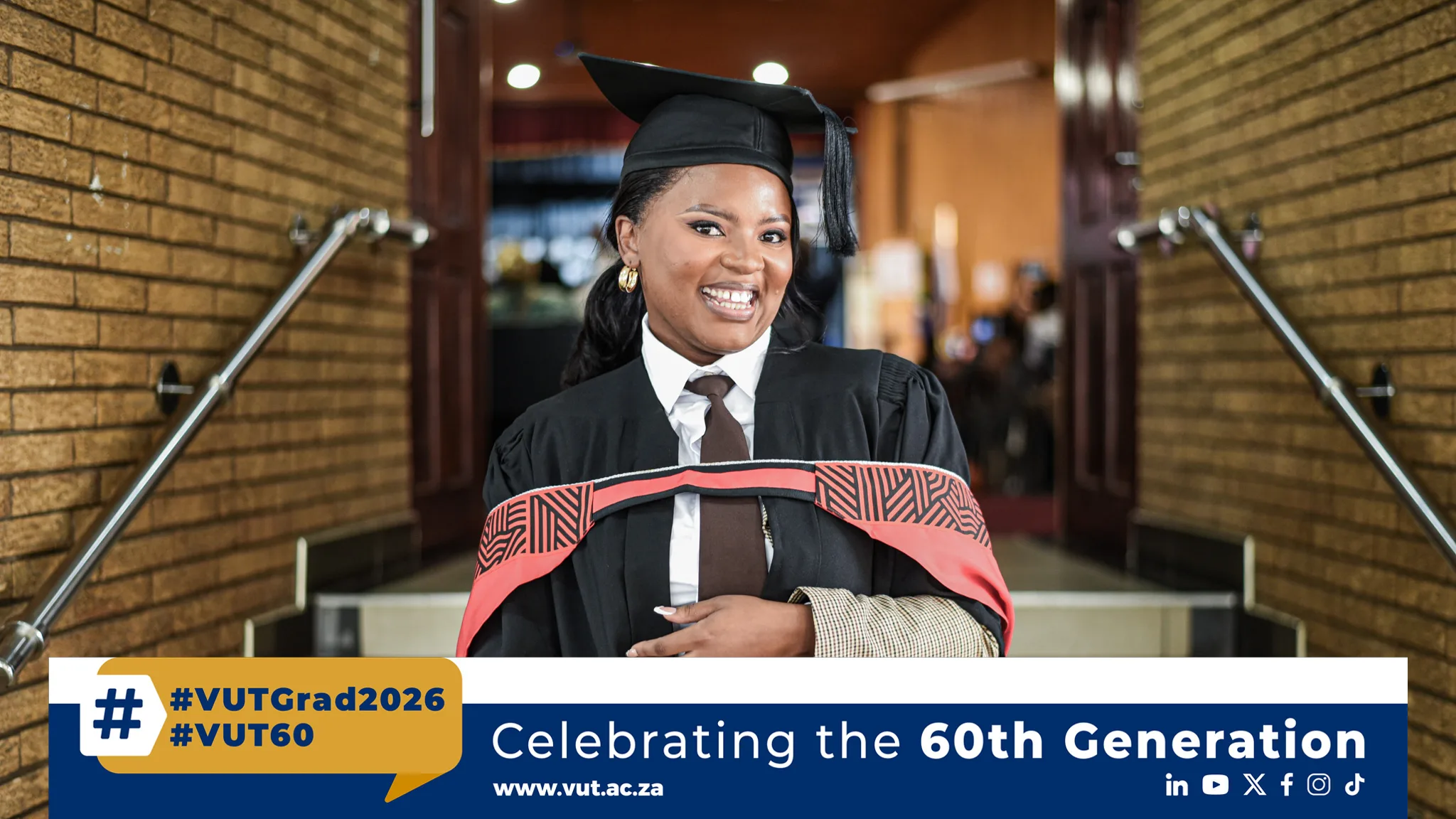 Graduate in cap and gown smiling at the camera in a hallway, banner partially visible reads '#VUTGrad2026' and 'Celebrating the 60th Generation'