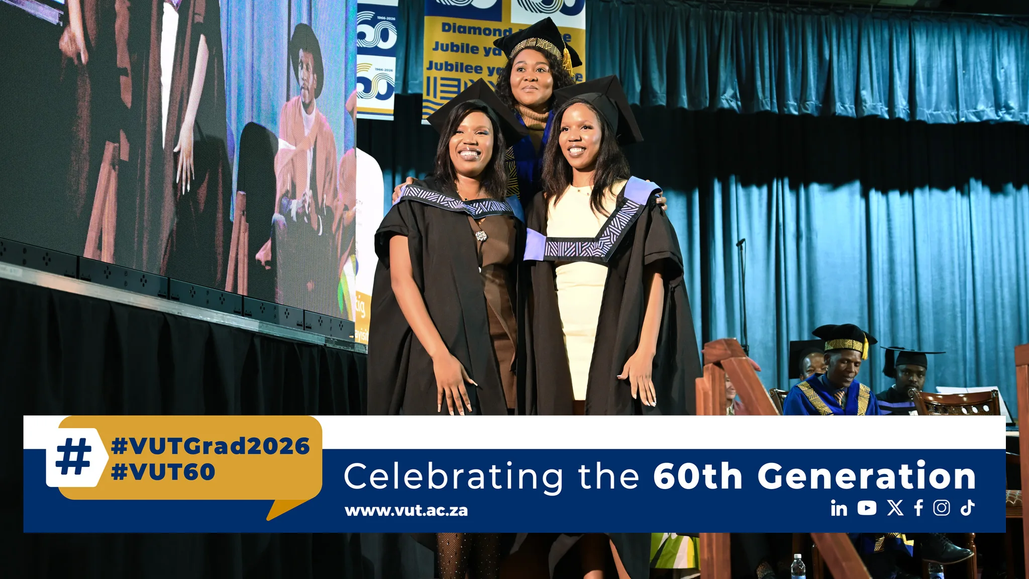 Twins – Vaal University of Technology Two graduates in black gowns and mortarboards pose on a stage, smiling, with a blue curtain backdrop and a banner that reads #VUTGrad2026 and Celebrating the 60th Generation on the bottom edge.