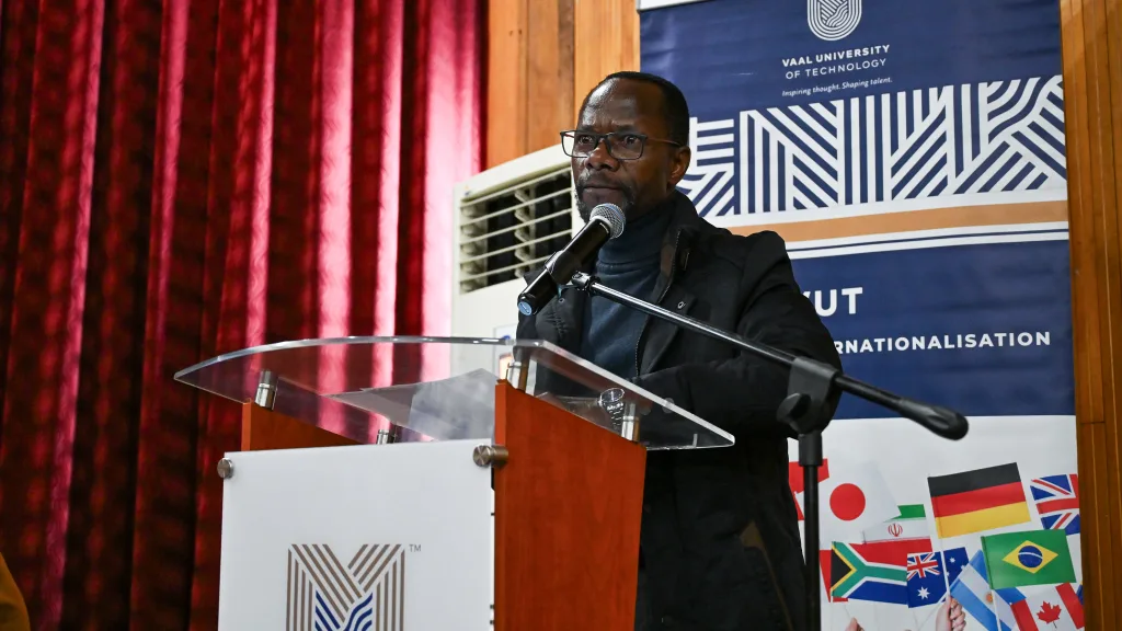 55264970631_d0724be16e_o – Vaal University of Technology Man at a podium giving a speech, microphone in hand, with a blue banner and international flag collage in the background.