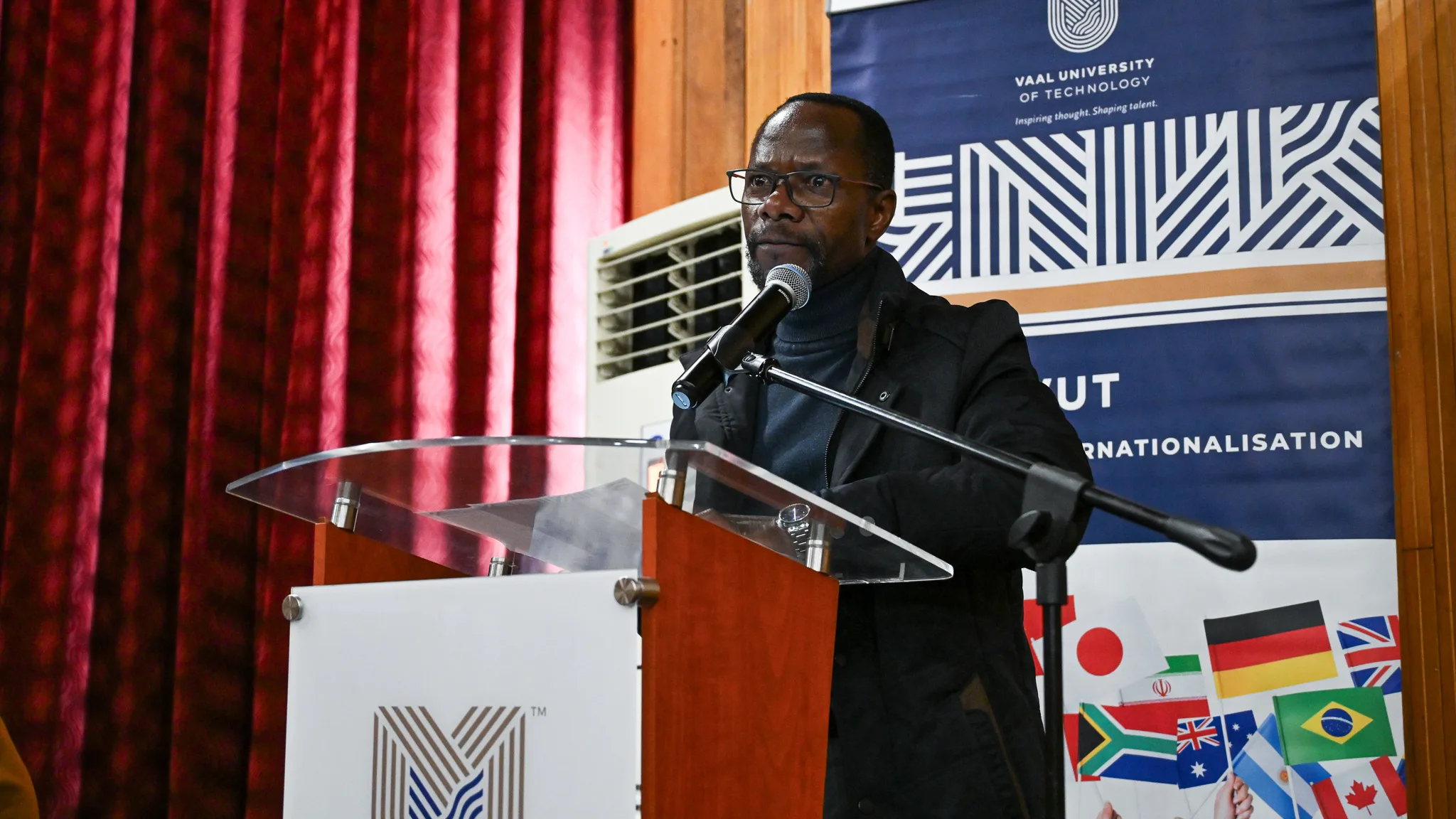 Man at a podium giving a speech, microphone in hand, with a blue banner and international flag collage in the background.