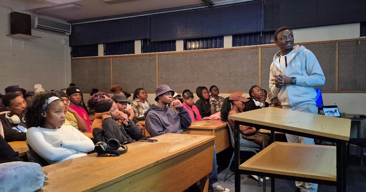 Fine art – Vaal University of Technology Instructor stands at a lectern addressing a classroom full of students seated at wooden benches.