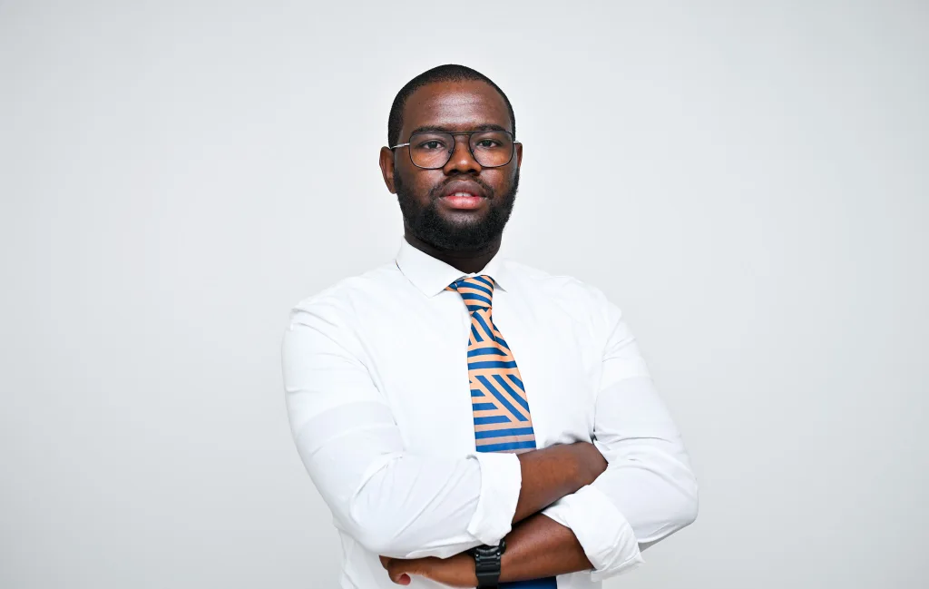 Portrait of a man with glasses and an orange-and-blue striped tie, arms crossed, facing the camera against a light gray background.