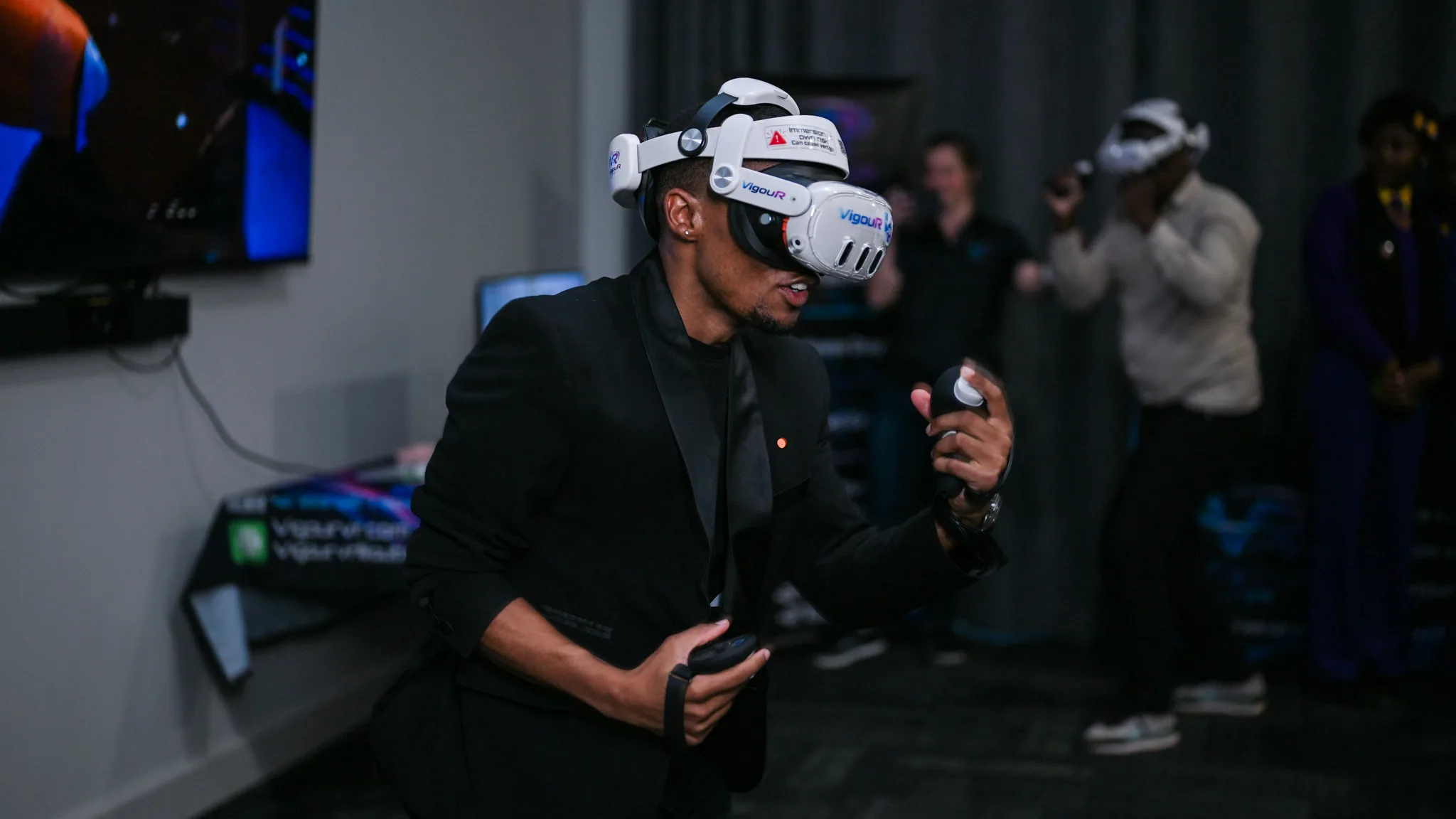 Meterverse – Vaal University of Technology – Vaal University of Technology Person wearing a white VR headset and black suit holds a controller during a dimly lit VR demonstration.