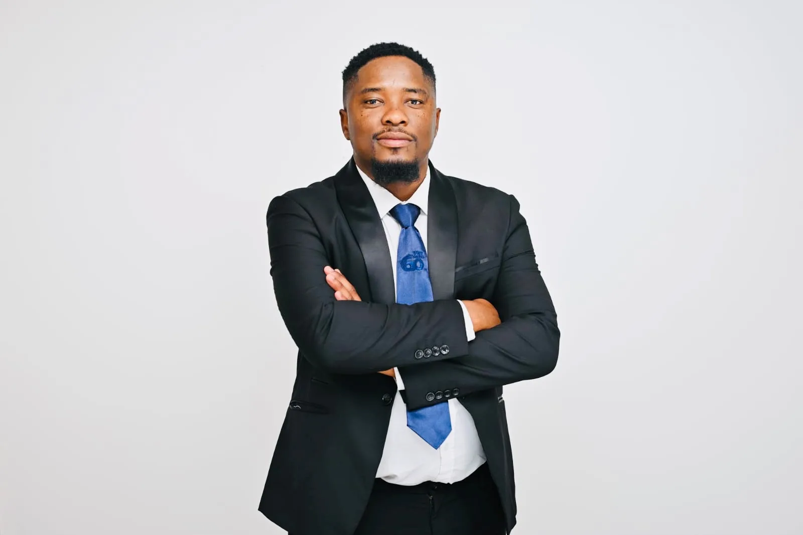 Professional headshot of a man in a black suit and blue tie with arms crossed against a light gray background.