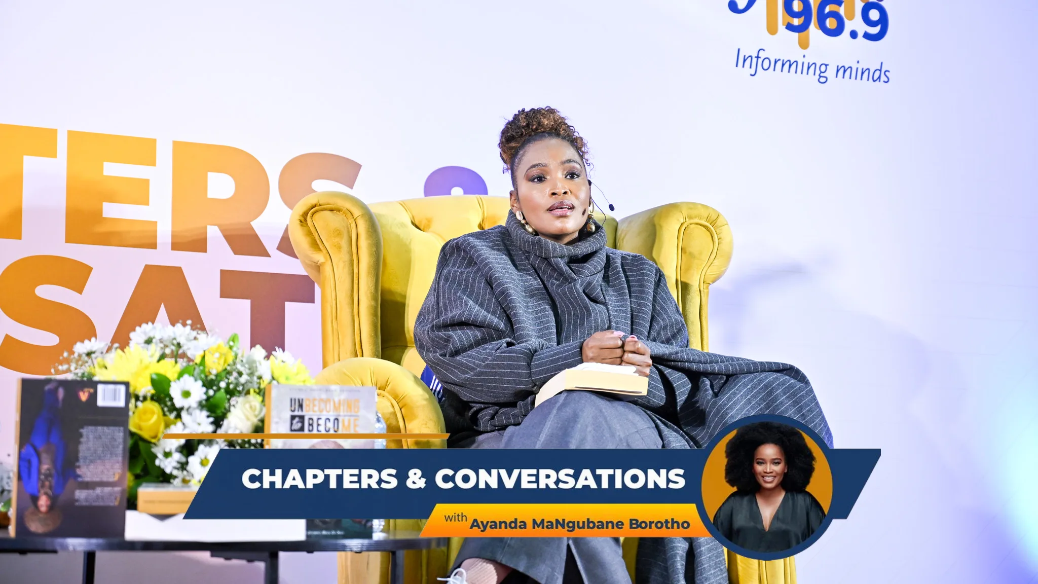 Ayanda MaNgubane Borotho-299 – Vaal University of Technology – Vaal University of Technology Woman seated in a yellow armchair on a stage, wearing a gray striped outfit and headset mic, speaking at a panel with a 'Chapters & Conversations' banner and books nearby