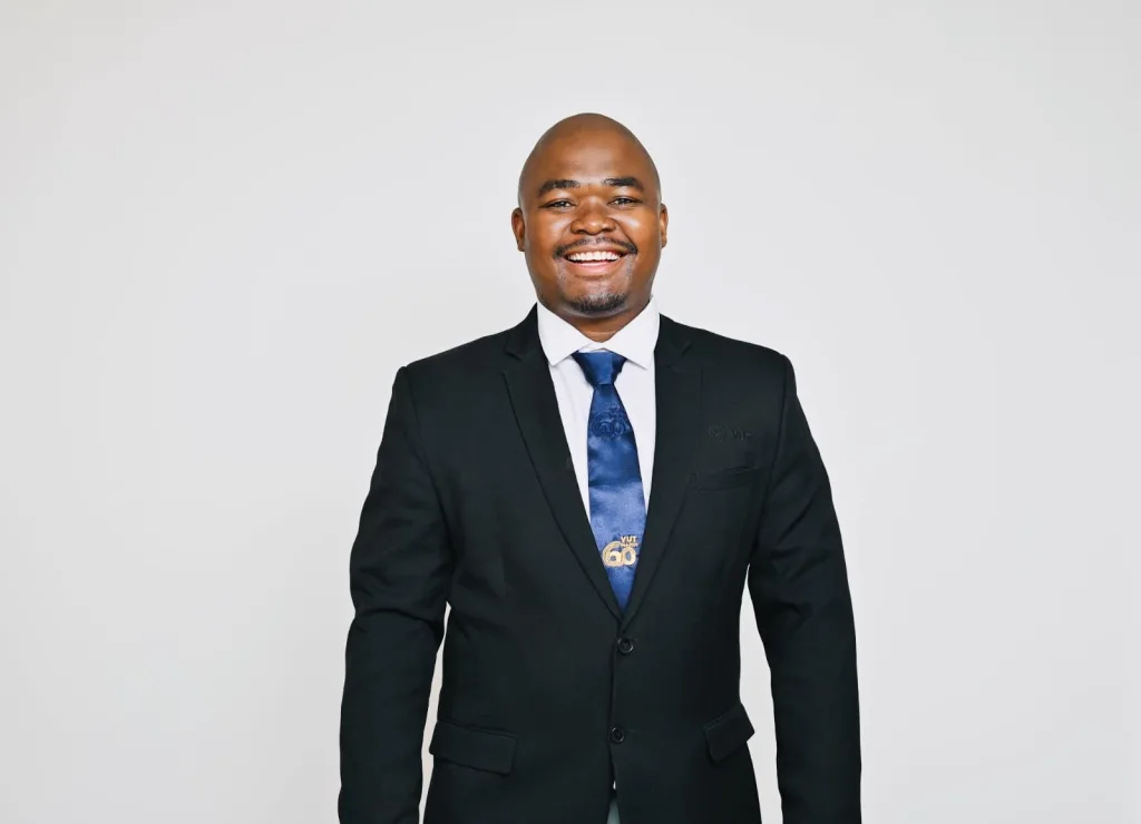 Smiling man in a black suit and blue tie against a neutral gray studio background.