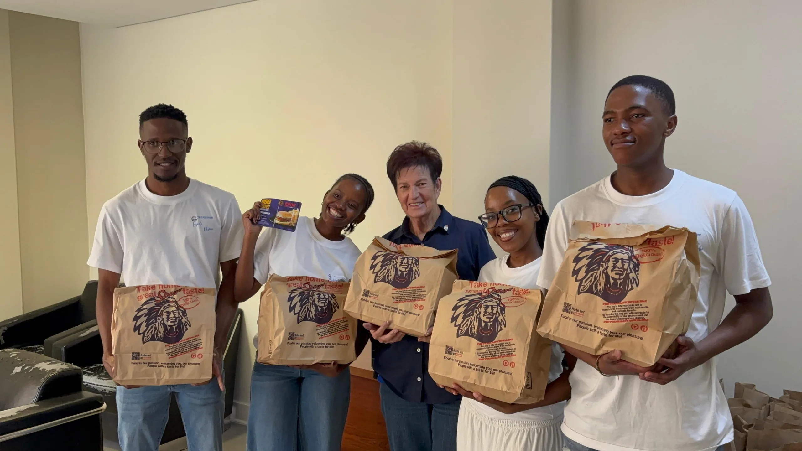 IMG-20260522-WA0269 1 – Vaal University of Technology – Vaal University of Technology Five volunteers indoors, smiling, each holding large brown paper bags with a lion logo (meal bags) for a food drive or donation event.