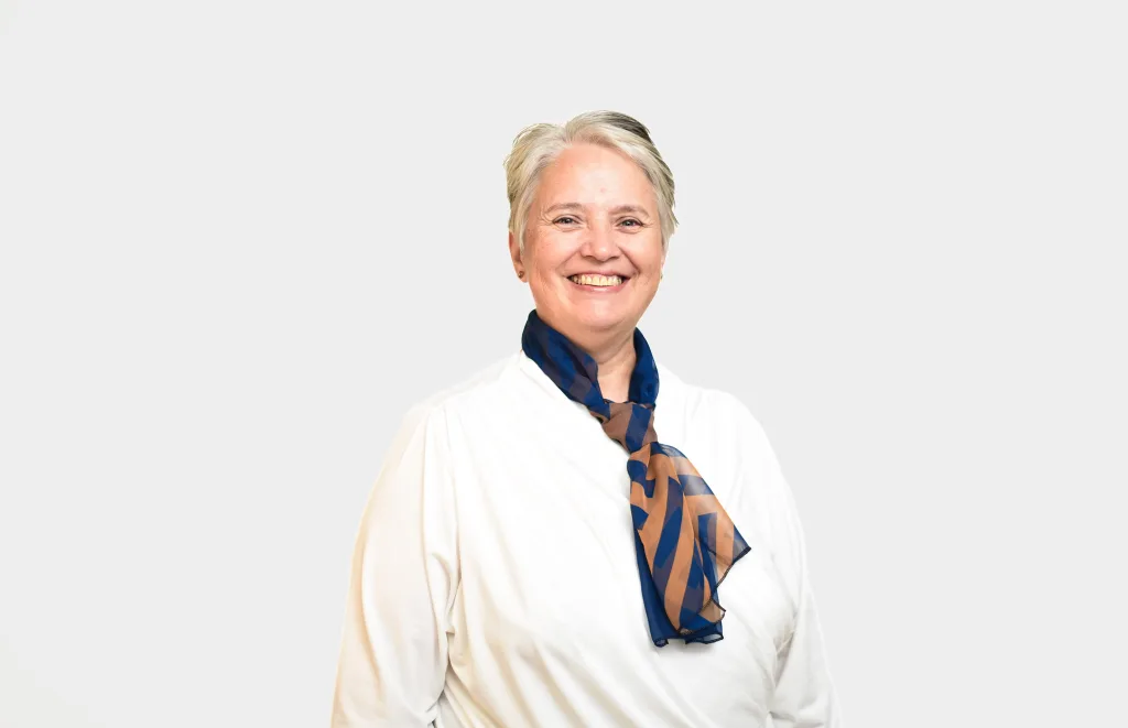 Smiling woman with short gray hair wearing a white top and blue-orange scarf against a light gray background.