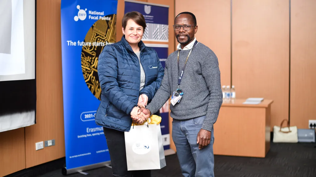 Ms Huba Boshoff – Vaal University of Technology Two people shaking hands at a formal event, smiling, with blue event banners in the background.