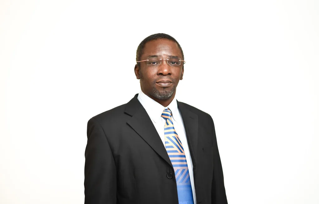 Portrait of a man in a black suit, blue striped tie, and glasses, looking at the camera against a white background.