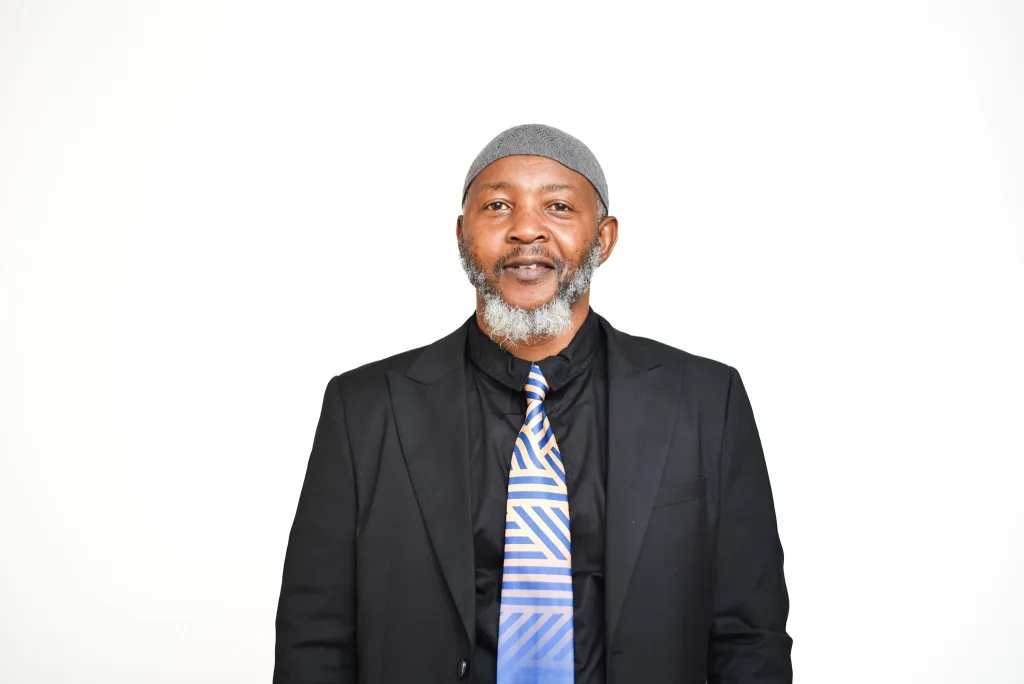 Portrait of a man in a dark suit and blue striped tie, wearing a gray knit cap, against a white background.