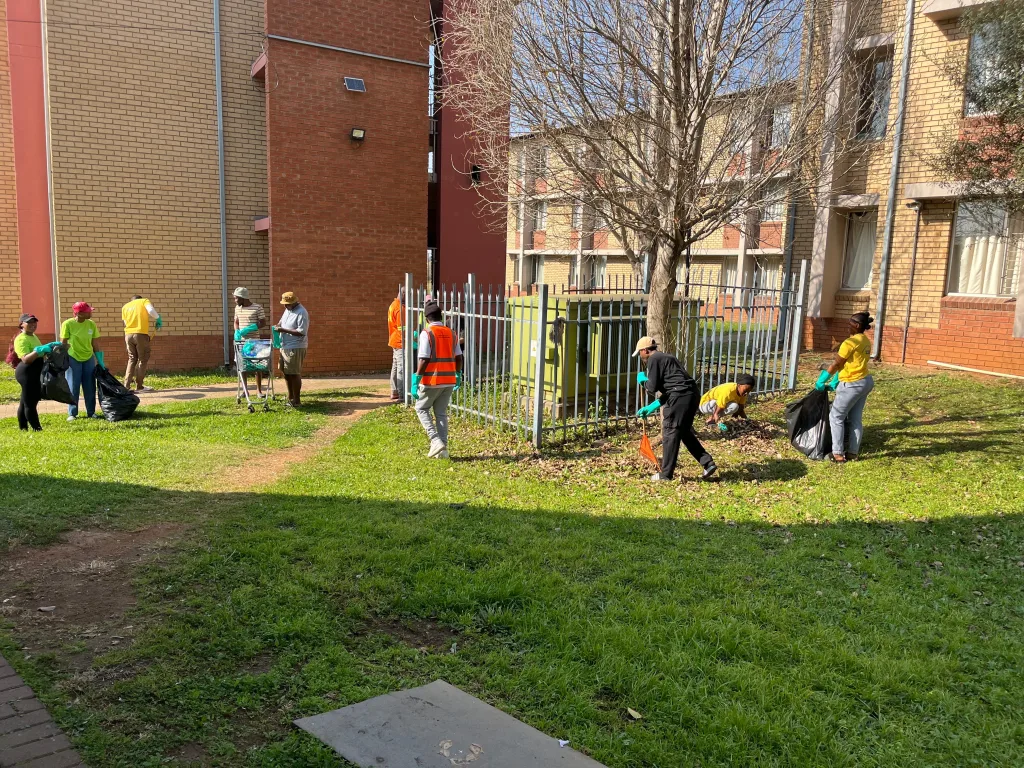 Residence students lead successful clean-up campaign to promote healthy living – Vaal University of Technology People wearing gloves and safety vests pick up litter around a courtyard, filling black bags near a metal fence and brick buildings.