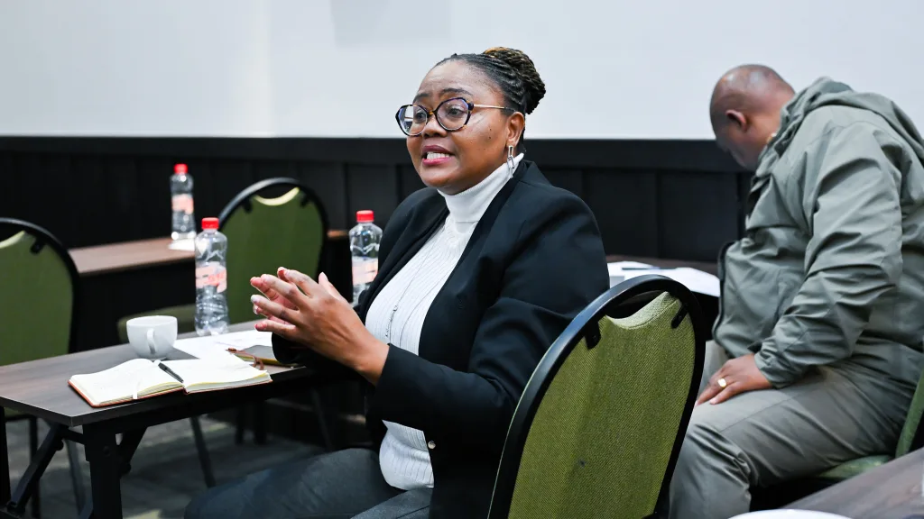 Teaching Advancements at Universities-05 – Vaal University of Technology Woman in glasses and blazer speaking at a meeting table, hands gesturing, with notebooks and water bottles nearby.