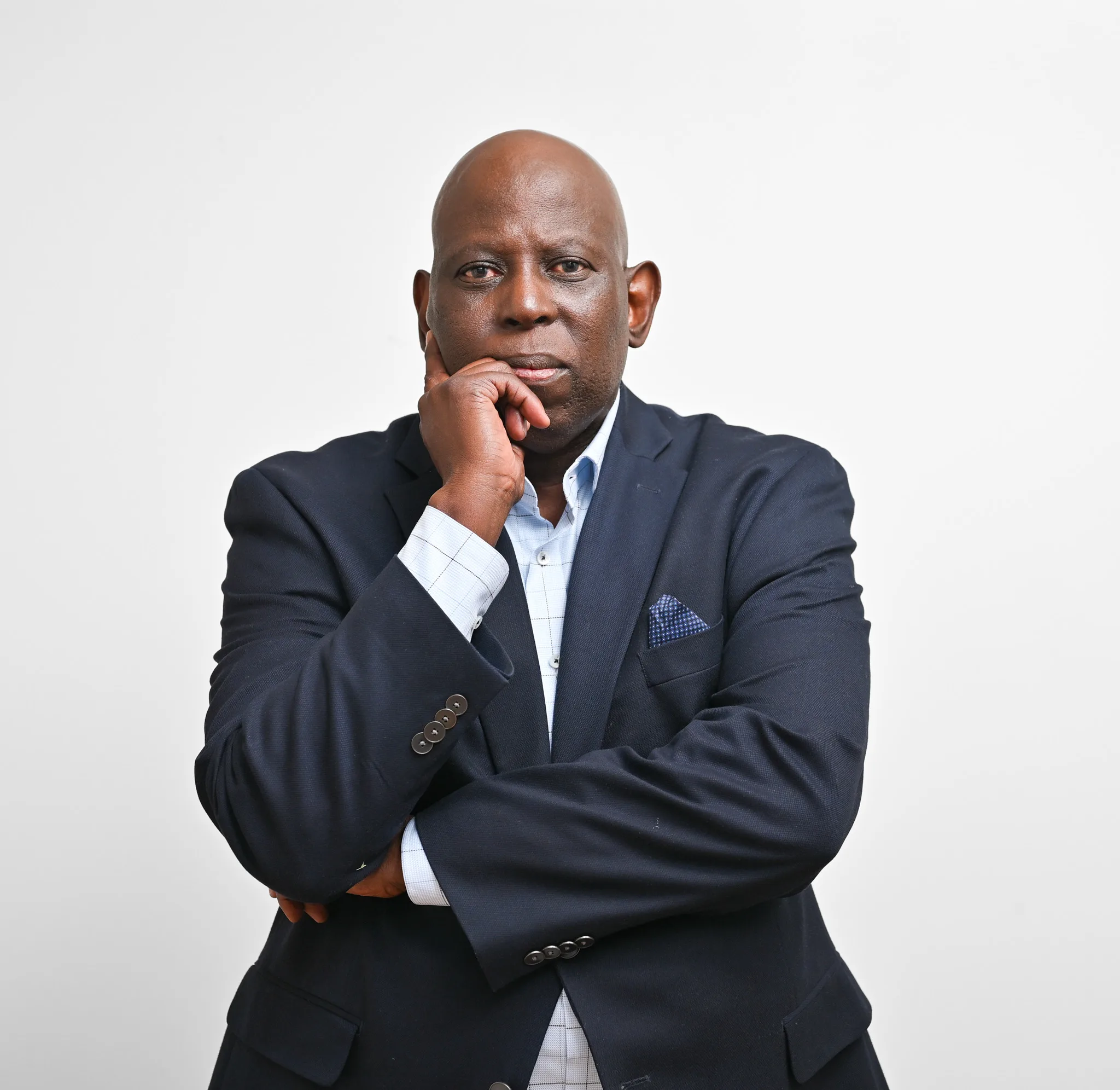 Thoughtful man in a navy suit with a light blue shirt, resting his chin on his hand against a plain white background.