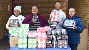 sanitary towels drive – Vaal University of Technology – Vaal University of Technology Four women stand behind a table piled with donated sanitary products (Kotex packs) and smile at the camera, with a brick wall background on either side.