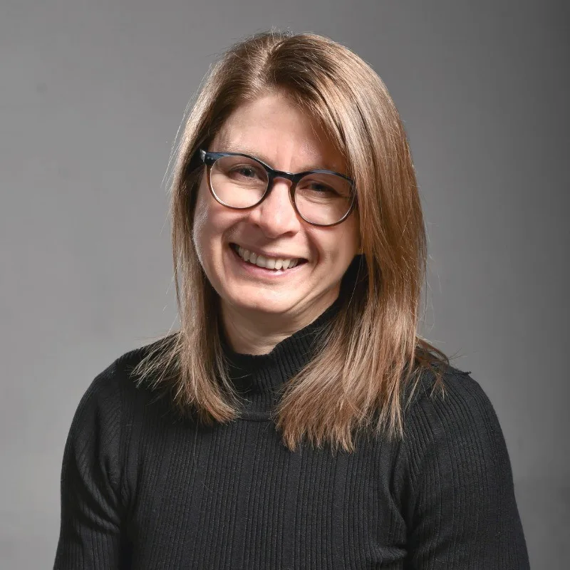 Portrait of a smiling woman with shoulder-length light brown hair and glasses wearing a black turtleneck, against a gray studio backdrop.