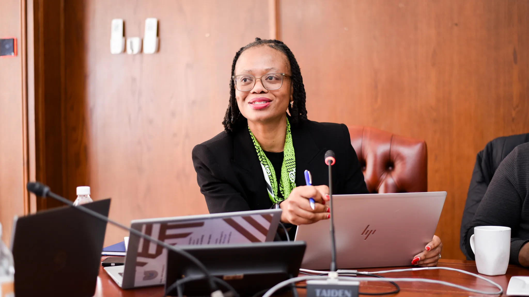Female academic leaders fellowship – Vaal University of Technology – Vaal University of Technology Professional woman with glasses and a green scarf speaking at a conference table with laptops and microphones in front of her.