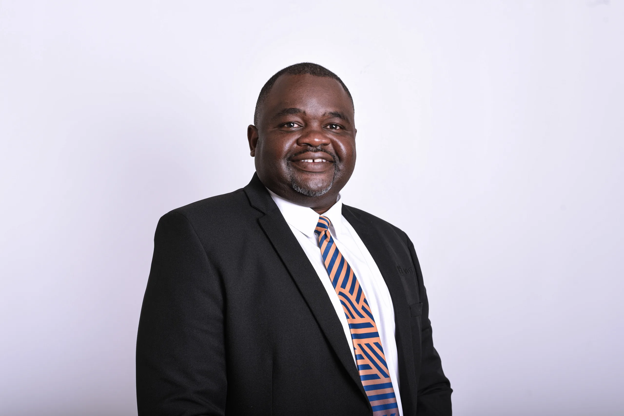 Smiling man in a dark suit and striped orange-blue tie against a light studio backdrop.