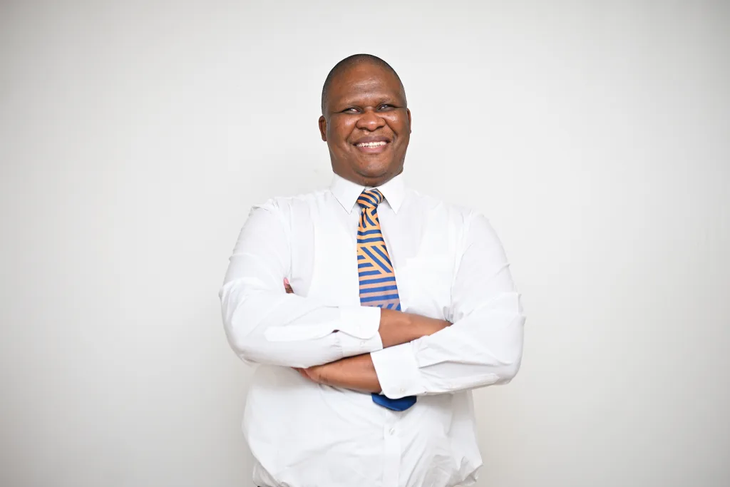 Smiling man in a white shirt and orange-blue striped tie, arms crossed, against a neutral background.