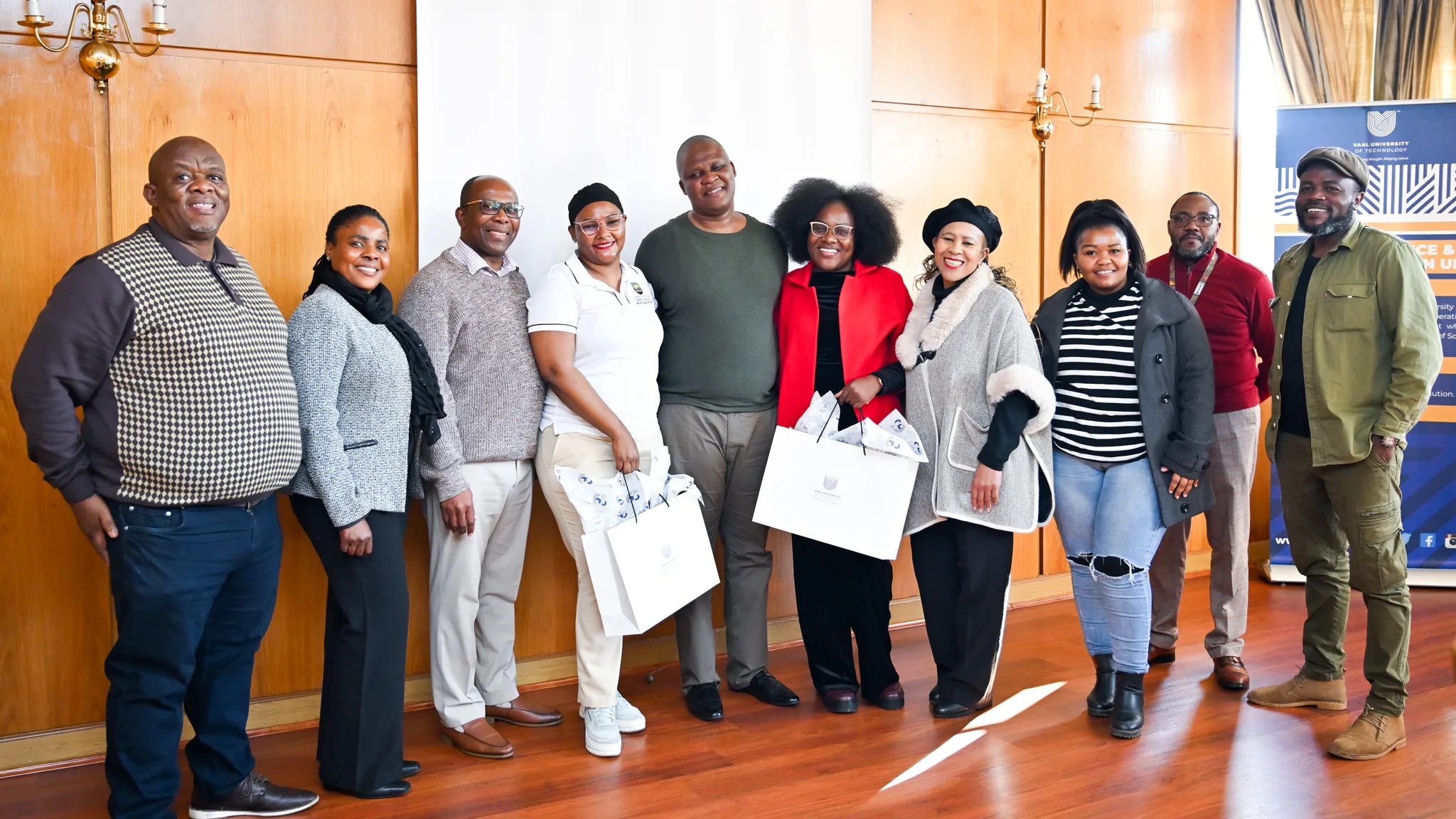 SJT Benchmarking Meeting with UFH-50 – Vaal University of Technology – Vaal University of Technology Group of diverse adults posing for a photo in a room with wooden panels; several hold white gift bags and smile at the camera, suggesting a ceremony or event.
