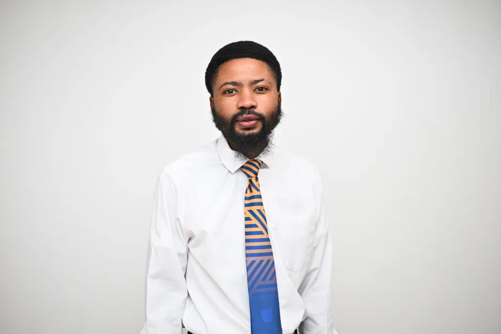 Portrait of a man with a full beard wearing a white dress shirt and an orange-and-blue striped tie against a pale background.