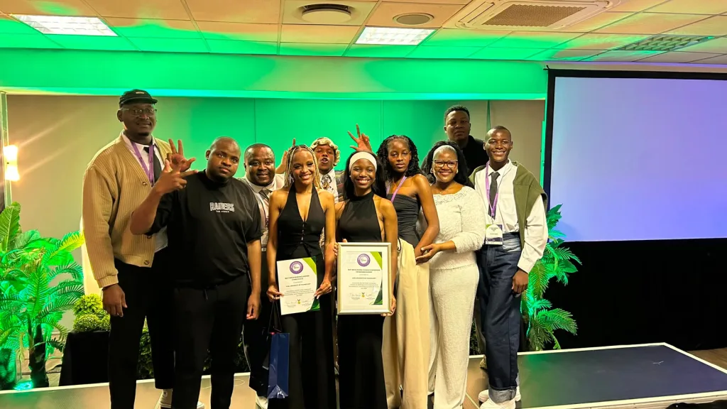 VUT Green Campus Initiative wins National Sustainability Award at Green Campuses Conference 2026 – Vaal University of Technology Group of people at an awards event posing for a photo; two women in black dresses hold certificates, others smile on a stage with green lighting and a projector screen in the background