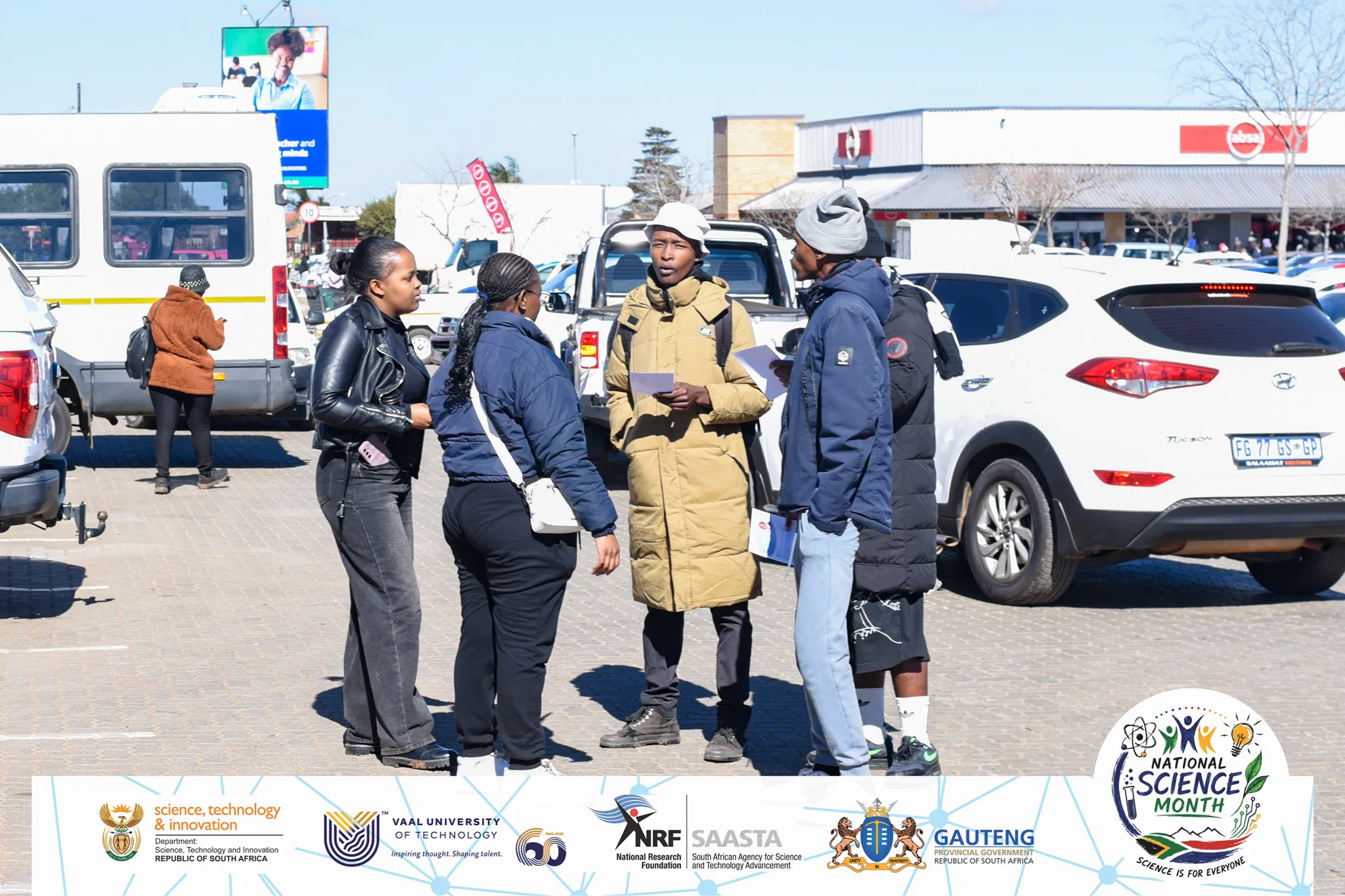 VUT takes the National Science Month campaign to the community 4 – Vaal University of Technology – Vaal University of Technology Group of young adults in jackets conversing in a sunny parking lot during an outdoor science event, with banners and cars in the background.