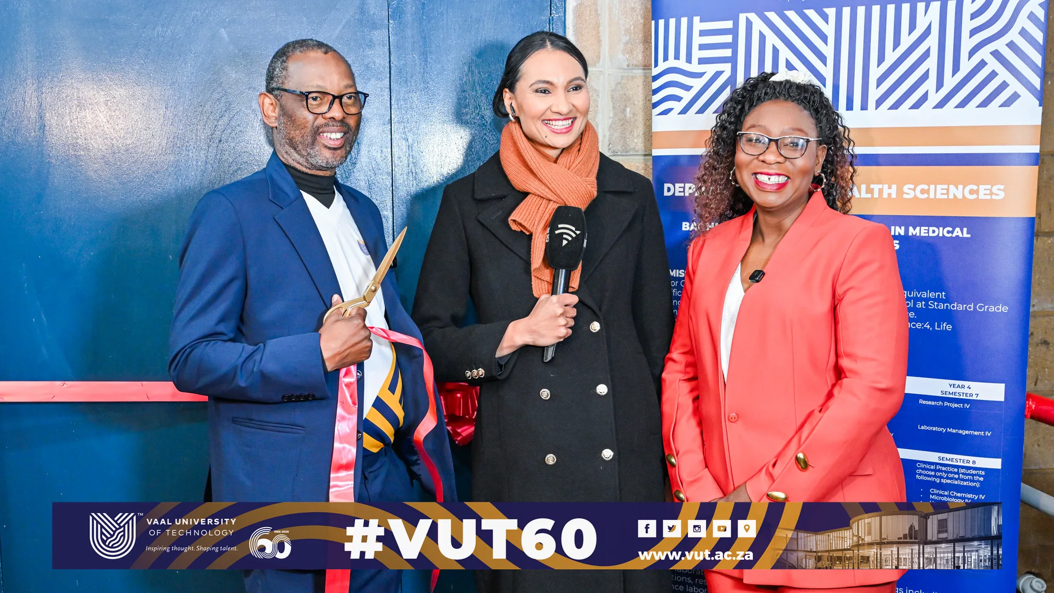 Three smiling presenters at a ribbon-cutting event in front of a VUT banner, with one man holding scissors and a ribbon while two women pose with a microphone.