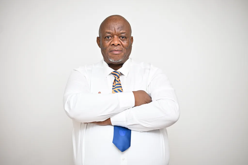 Portrait of a middle-aged man with arms crossed, wearing a white shirt and a blue-and-orange striped tie, against a plain light background.