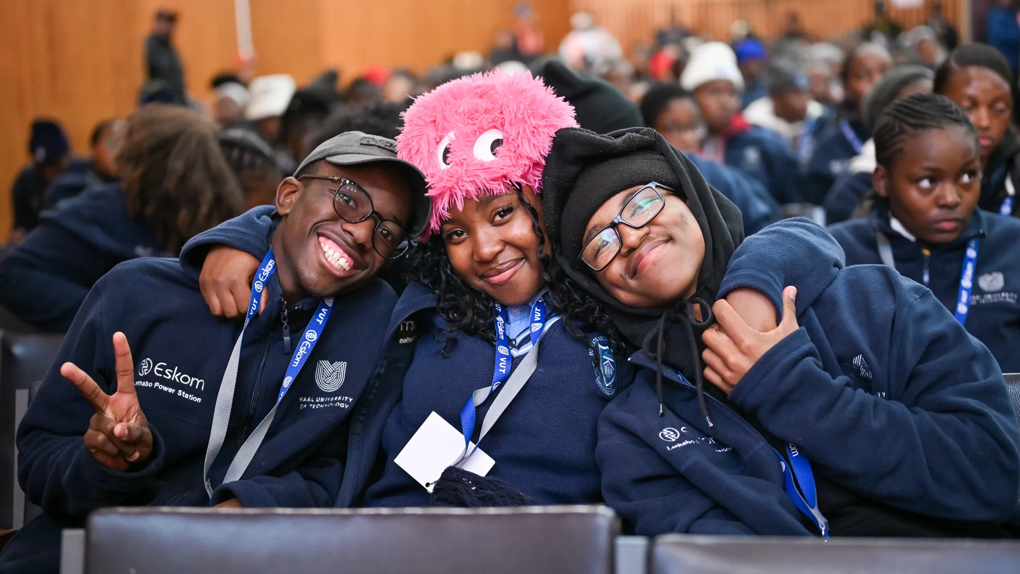 Three smiling students in navy hoodies with lanyards pose together with arms around each other at a crowded indoor event; the center student wears a pink fluffy hat.
