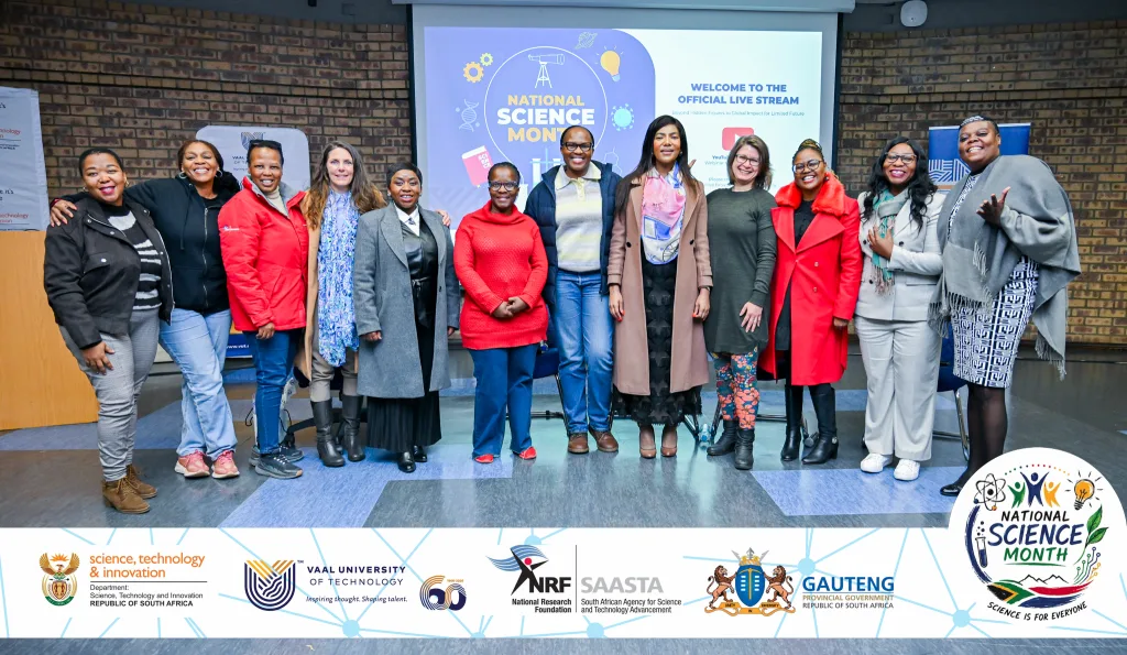 Womens Dialogue 2 – Vaal University of Technology Diverse group of adults posing for a photo at a National Science Month event, with banners and a projection screen behind.