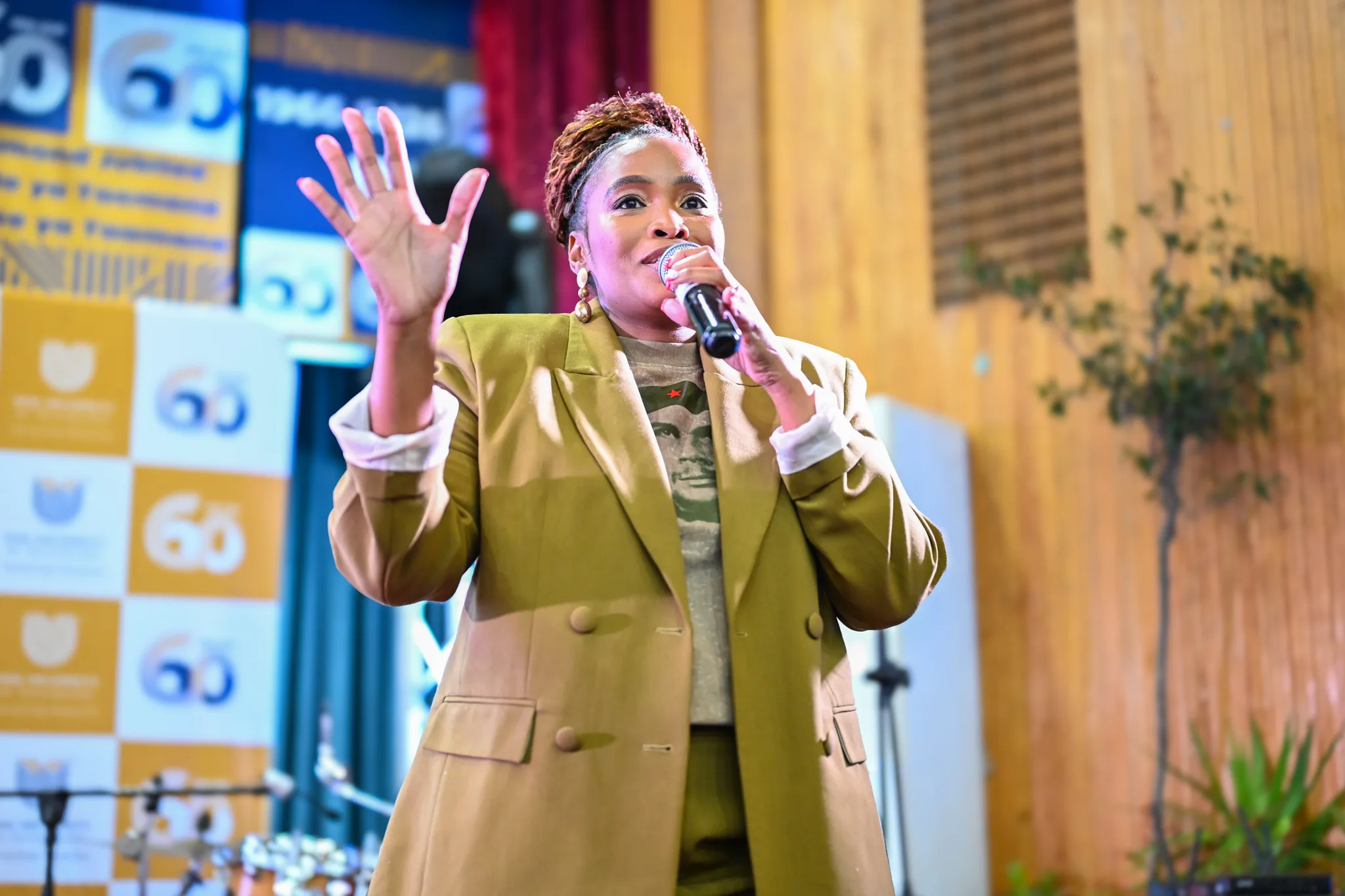 Woman singing into a microphone on stage, wearing a tan blazer and camo shirt, raising her left hand as she performs.