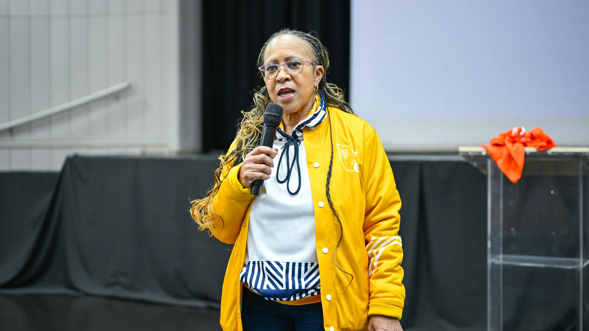 Capacity building strengthens partnerships for safer students residences at VUT – Vaal University of Technology – Vaal University of Technology Woman in a bright yellow jacket talks into a handheld microphone on a stage, wearing glasses and a striped scarf.