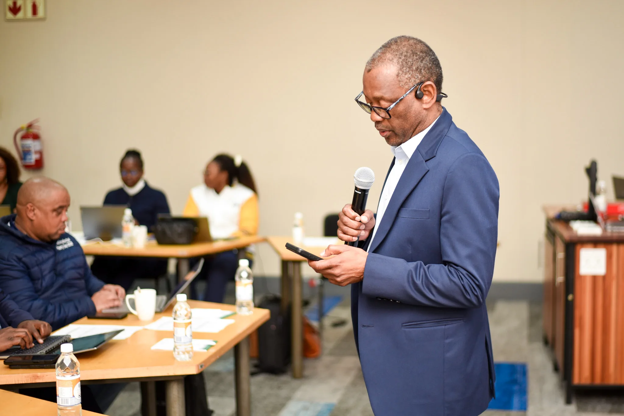 Employee Onboarding-22 – Vaal University of Technology – Vaal University of Technology Man in a blue suit speaks into a handheld microphone in a meeting room, others seated at tables with laptops and bottles nearby.