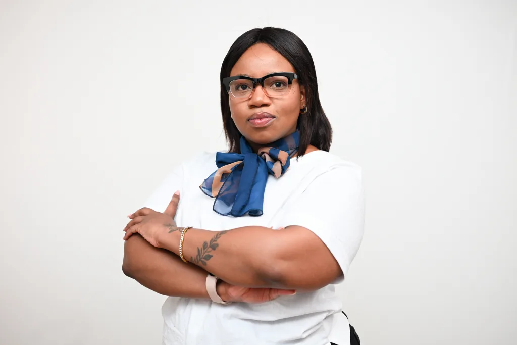 Woman with black hair and glasses, wearing a blue patterned scarf and white shirt, arms crossed against a light background.