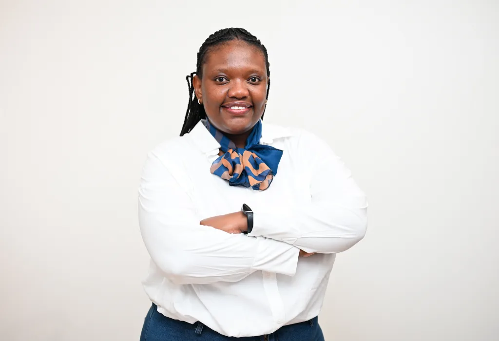 Smiling woman with braided hair, wearing a white shirt and blue scarf, arms crossed in front of a light background.