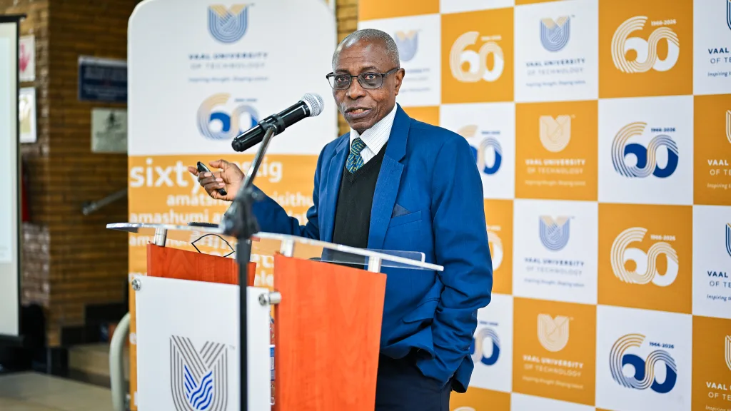 Prof James Darkwa – Vaal University of Technology Man in a blue suit and glasses speaks at a podium with a microphone, backed by orange-and-white branded banners.