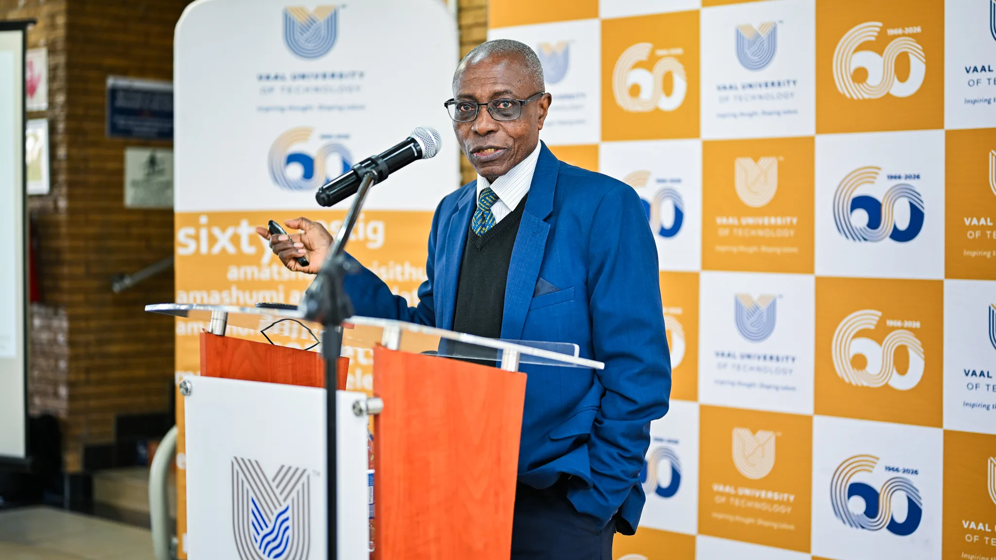 Prof James Darkwa – Vaal University of Technology – Vaal University of Technology Man in a blue suit and glasses speaks at a podium with a microphone, backed by orange-and-white branded banners.