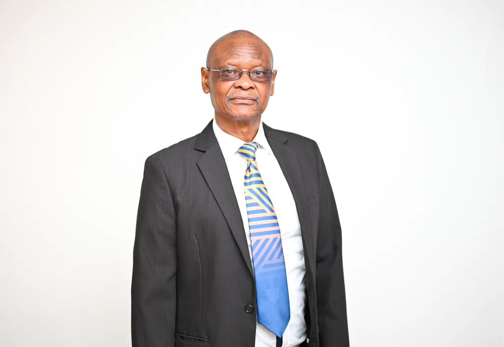 Man in a dark suit and striped blue tie, wearing glasses, standing against a light background with a neutral expression.