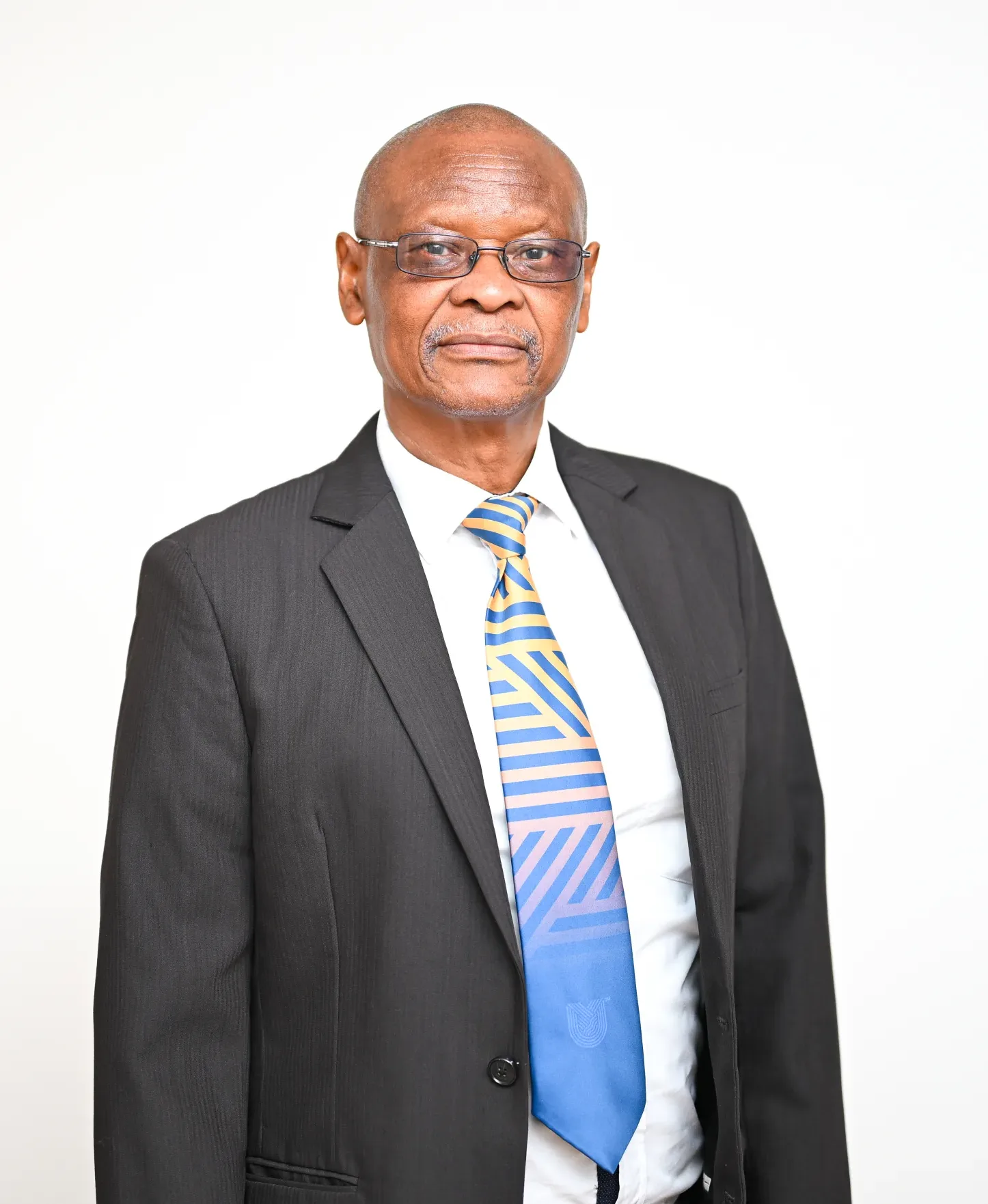 Portrait of an older man in a dark suit, white shirt, and striped blue-yellow tie, standing against a light background with a neutral expression, wearing glasses.