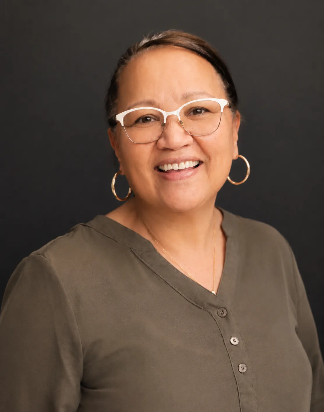 VUTs Shirene Klink earns prestigious RMP designation – Vaal University of Technology – Vaal University of Technology Smiling woman with white glasses and gold hoop earrings, wearing an olive blouse against a dark backdrop.