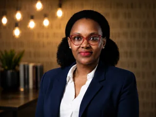 Portrait of a professional woman wearing a navy blazer and white shirt, with red glasses, in a modern office setting.