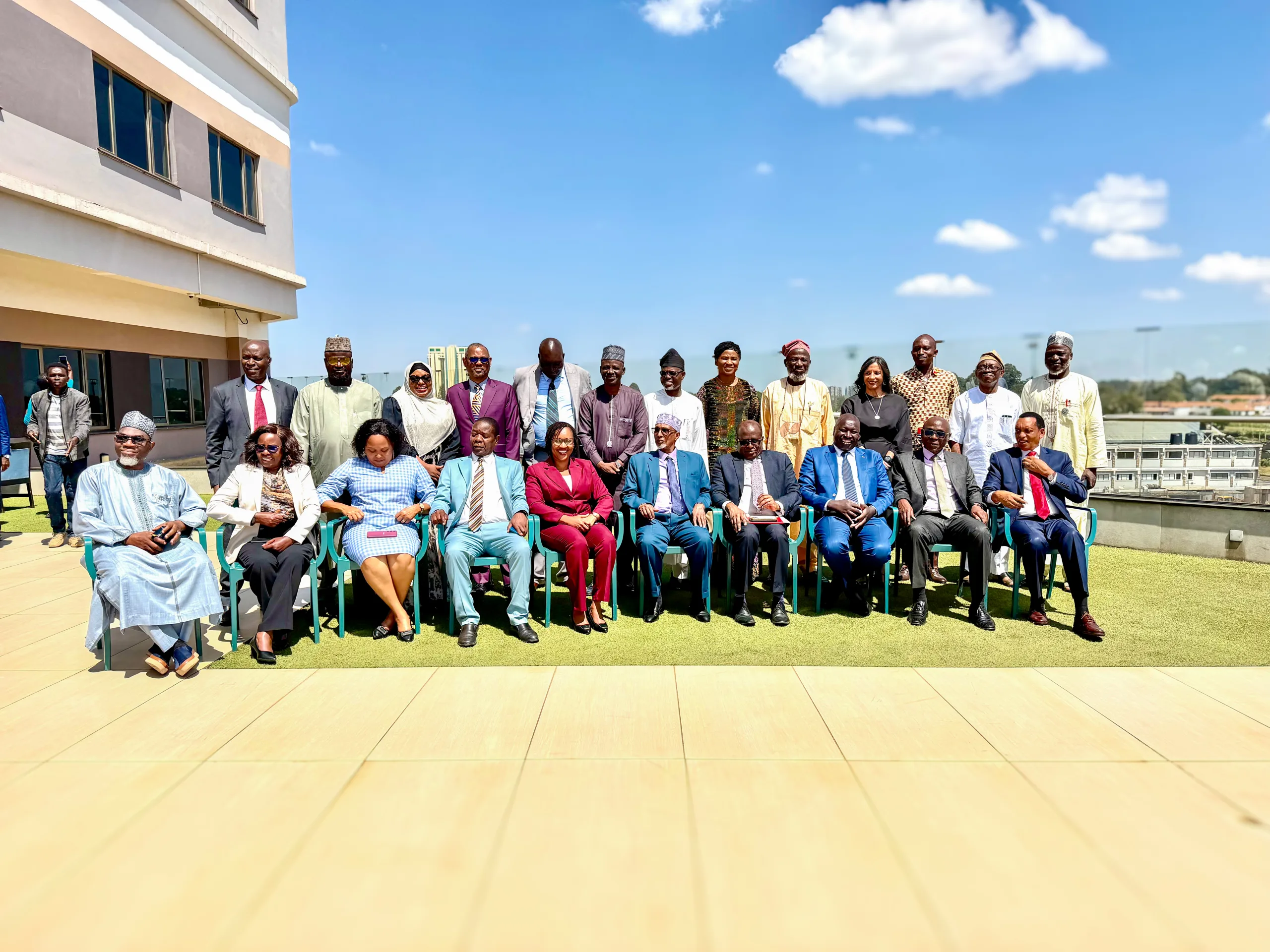 Large group of people in formal and traditional clothing pose for a group photo on an outdoor rooftop terrace with a blue sky and a building to the left.
