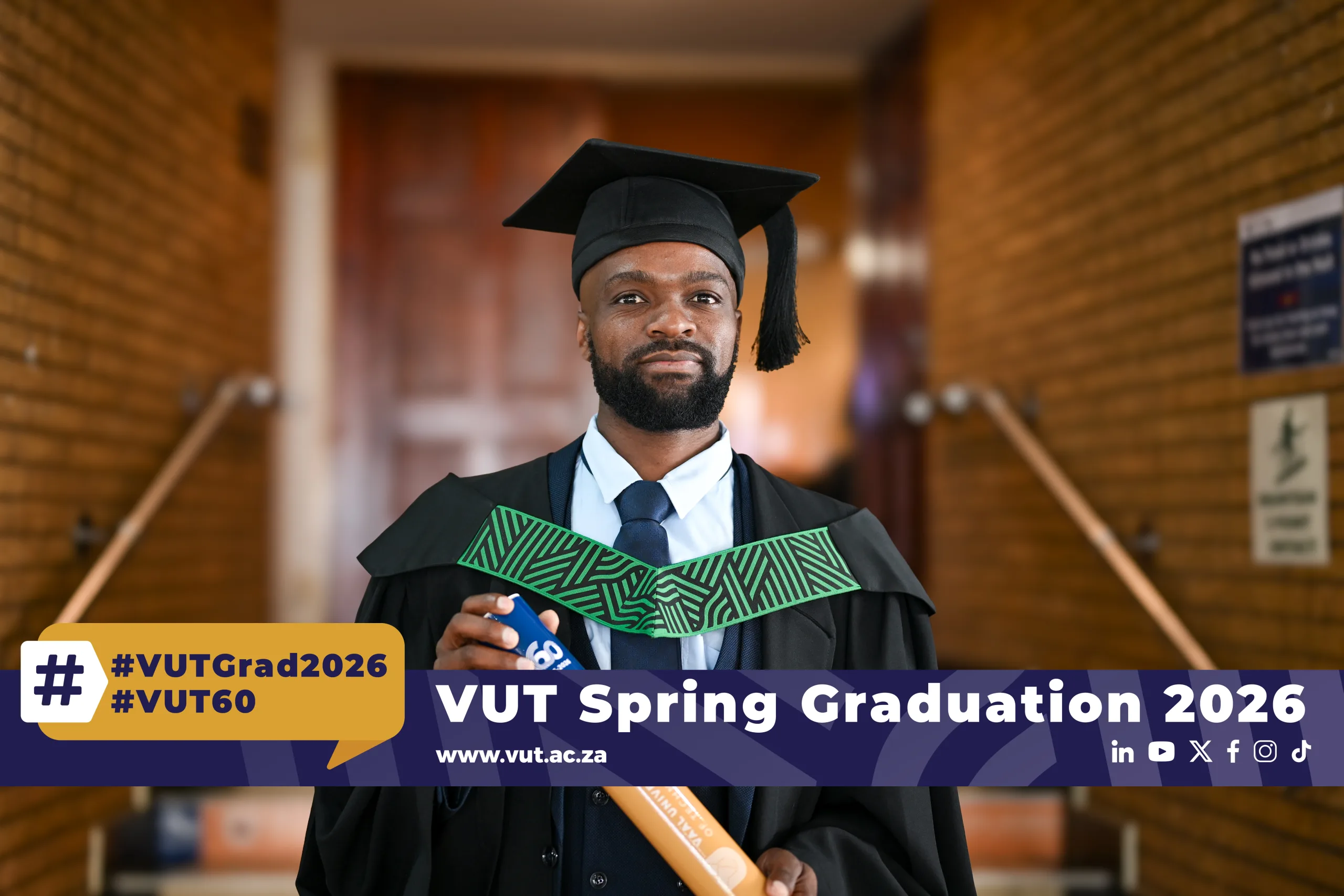 Graduate in cap and gown holding a diploma in a campus hallway with a banner reading 'VUT Spring Graduation 2026'