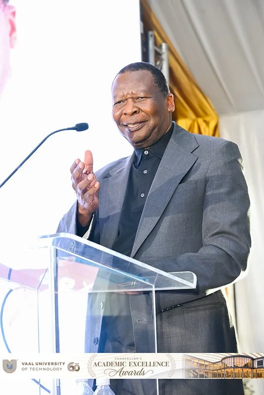 A man in a gray suit speaking at a clear podium with a microphone on stage during an awards ceremony.