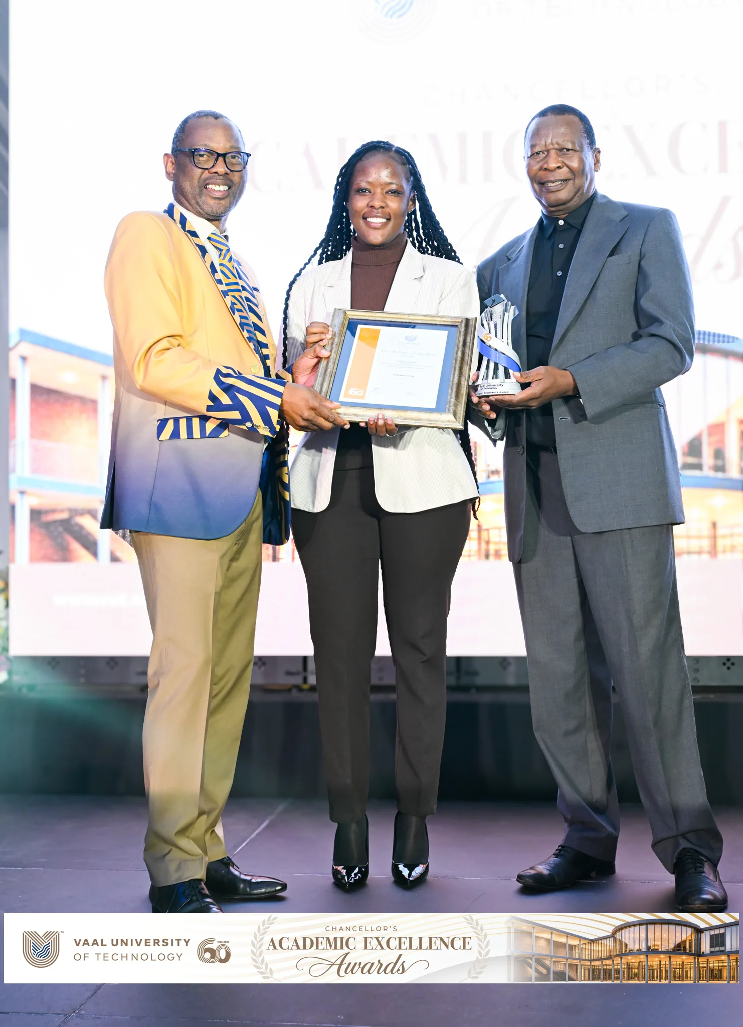 Three award recipients on a stage: a smiling woman in the center holds a framed certificate, flanked by two men in suits; one holds a trophy. The event appears to be an Academic Excellence Awards ceremony at a university.