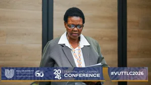 Ms Njo Makapela-Cikizwa – Vaal University of Technology – Vaal University of Technology African American woman in a gray blazer and glasses reads papers at a conference podium.